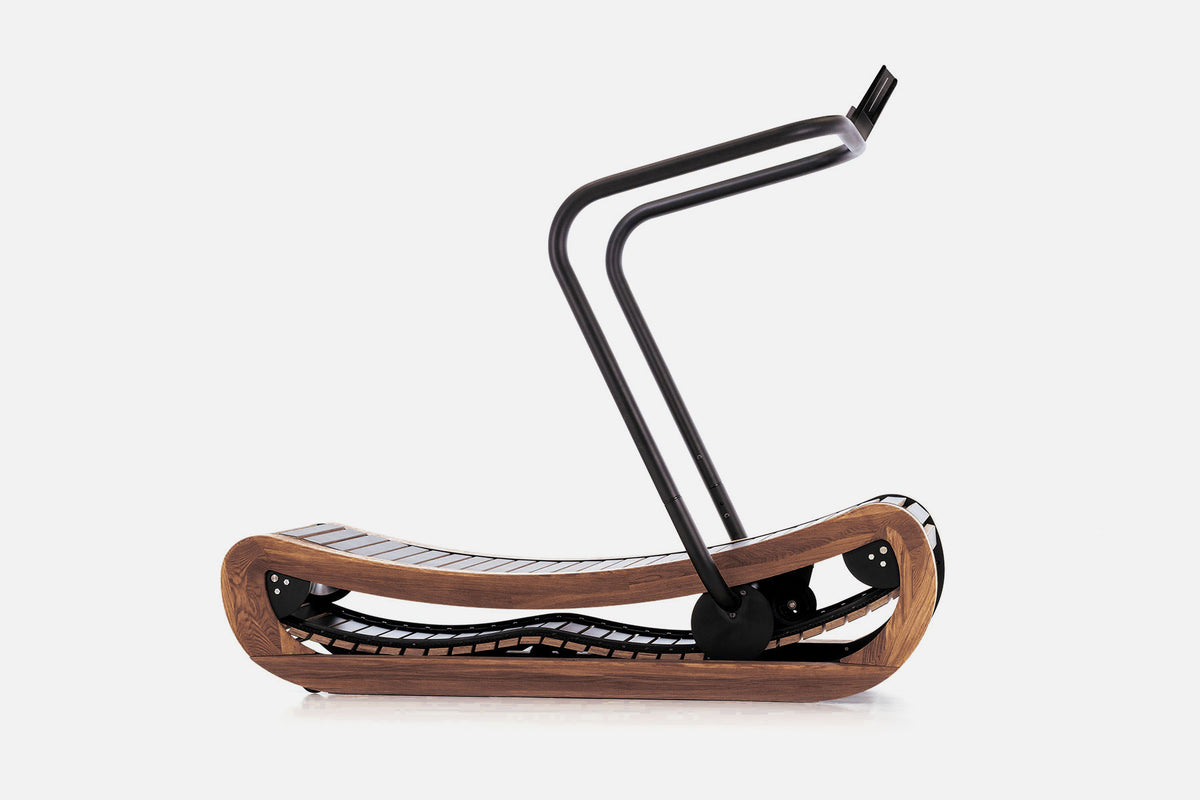 Side view of a curved manual treadmill with a medium-brown wood frame, black slats, and a raised black handlebar