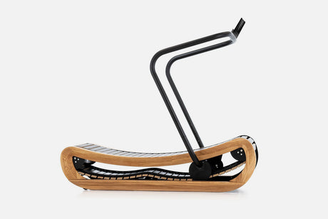 Side view of a curved manual treadmill with a pale wood frame, black slats, and a raised black handlebar